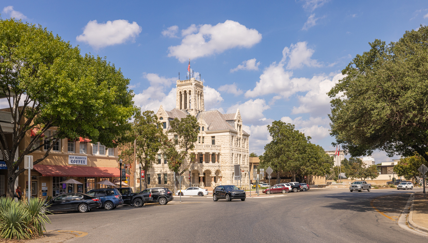 Downtown street scene with historic courthouse building, parked cars, and leafy trees in New Braunfels, TX.